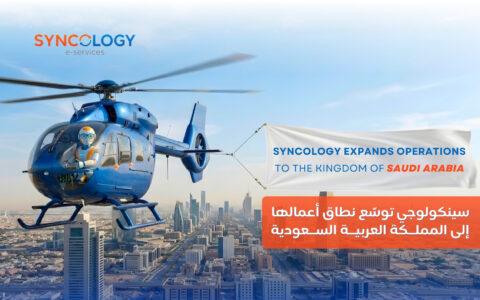 A blue helicopter piloted by a robot flying over a Saudi Arabian city skyline, pulling a banner that reads Syncology expands operations to the Kingdom of Saudi Arabia.