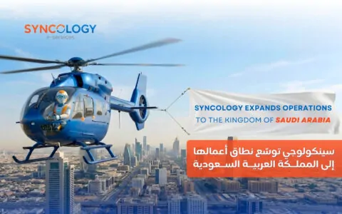 A blue helicopter piloted by a robot flying over a Saudi Arabian city skyline, pulling a banner that reads Syncology expands operations to the Kingdom of Saudi Arabia.